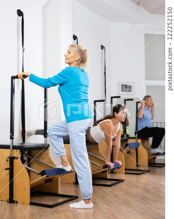 Positive mature female using Wanda Pilates chair equipment and making exercises for tone in fitness center Positive mature female using Wanda Pilates chair equipment and making exercises for tone in fitness center 122252150