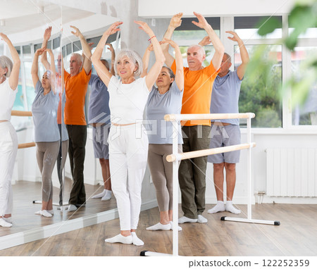 Active mature woman practicing fifth position holding by ballet barre together with others 122252359