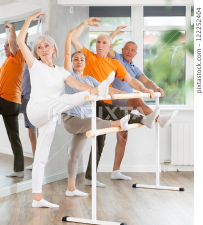 Active mature woman practicing leg stretching holding by ballet barre together with others 122252404