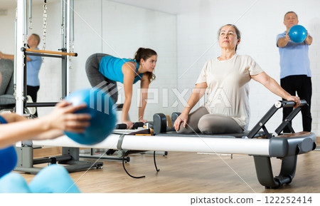 Serious mature female exercising and using Pilates performer bed in rehabilitation center Serious mature female exercising and using Pilates performer bed in rehabilitation center 122252414