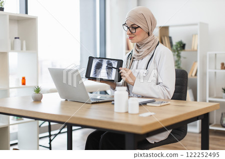 Muslim female doctor having video call with patient showing lung x-ray. Middle-aged woman in medical office using technology for telemedicine consultation. Muslim female doctor having video call with patient showing lung x-ray. Middle-aged woman in medical office using technology for telemedicine consultation. 122252495