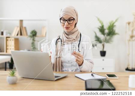 Middle Eastern female doctor in hijab conducts video call, offering patient treatment with pills, using laptop in modern medical office. 122252518