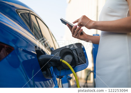 Woman is connecting an electric vehicle to a charger and checking her smartphone at a station. Woman is connecting an electric vehicle to a charger and checking her smartphone at a station. 122253094