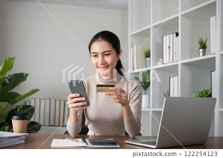 Beautiful young woman uses laptop to shop online, buys things and pays with credit card through phone online banking app. 122254123
