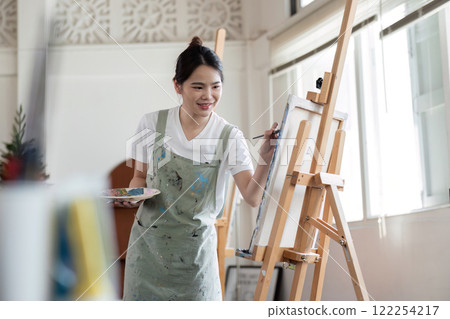 Young Asian female artist is intently painting acrylic paints on canvas in a painting studio. 122254217