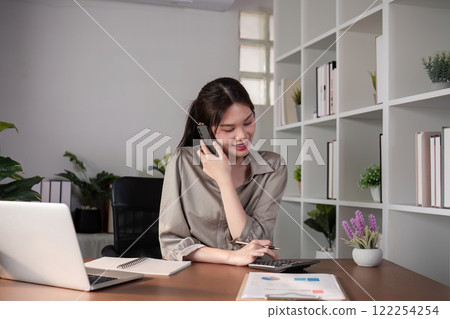 Young Asian business woman sits on the phone in an online business meeting using a laptop in a modern home office decorated with shady green plants. Young Asian business woman sits on the phone in an online business meeting using a laptop in a modern home office decorated with shady green plants. 122254254