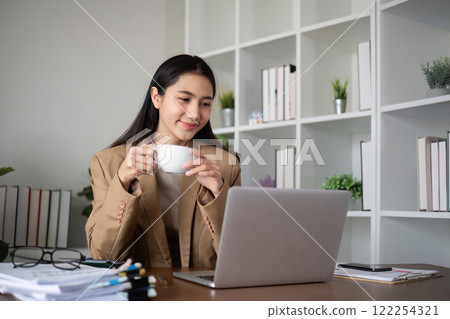 Young Asian business woman sits and works using a laptop in a modern office decorated with shady green plants. 122254321