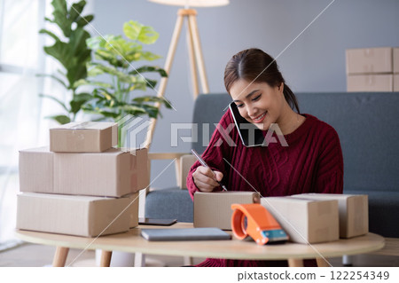 A small SME business owner receives product orders and writes shipping information on cardboard boxes in the home office to prepare for delivery. 122254349
