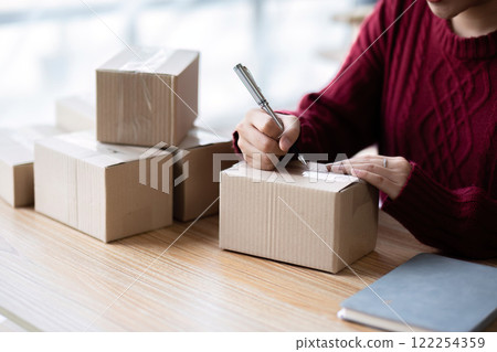 A small SME business owner receives product orders and writes shipping information on cardboard boxes in the home office to prepare for delivery. A small SME business owner receives product orders and writes shipping information on cardboard boxes in the home office to prepare for delivery. 122254359