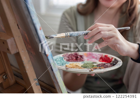 Young Asian artist woman holding a palette of colorful paints is intently painting acrylic paints on canvas in a painting studio. Young Asian artist woman holding a palette of colorful paints is intently painting acrylic paints on canvas in a painting studio. 122254391