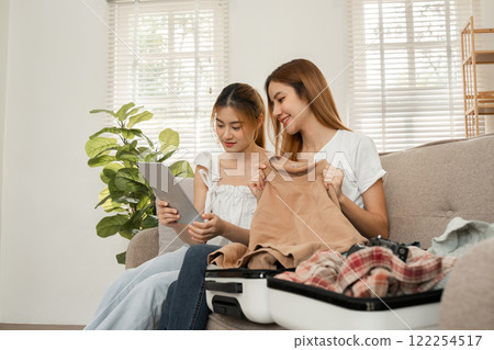 Two young female friends use a tablet to view travel information online for a vacation together. While packing your suitcase Two young female friends use a tablet to view travel information online for a vacation together. While packing your suitcase 122254517