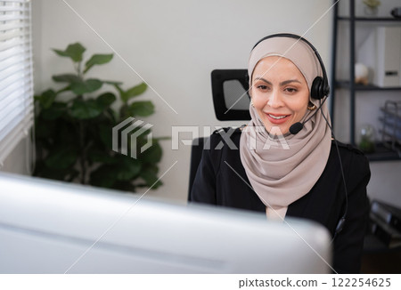 Call center worker, young Muslim woman wearing hijab, talking to customer on call phone on computer in customer service office Call center worker, young Muslim woman wearing hijab, talking to customer on call phone on computer in customer service office 122254625