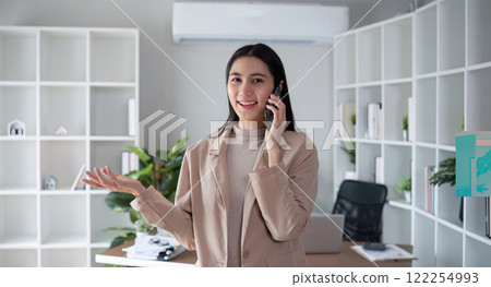 Young Asian business woman sits on the phone in an online business meeting using a laptop in a modern home office decorated with shady green plants. Young Asian business woman sits on the phone in an online business meeting using a laptop in a modern home office decorated with shady green plants. 122254993