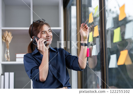Asian businesswoman talking in a work meeting on the phone, taking notes on sticky notes on the wall in the office Asian businesswoman talking in a work meeting on the phone, taking notes on sticky notes on the wall in the office 122255102