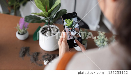 A young Asian woman uses her phone to take photos and enjoys planting plants in her garden to create a pleasant atmosphere in the house. 122255122