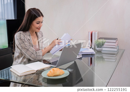 A middle-aged woman works from home at a dining room table with snacks and piles of documents on it. A middle-aged woman works from home at a dining room table with snacks and piles of documents on it. 122255191