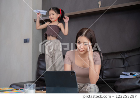 Stressed business mother sitting from home, taking notes and looking after her laptop with her cute daughter playing next to her. 122255287