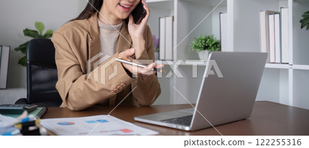 Young Asian business woman sits on the phone in an online business meeting using a laptop in a modern home office decorated with shady green plants. 122255316