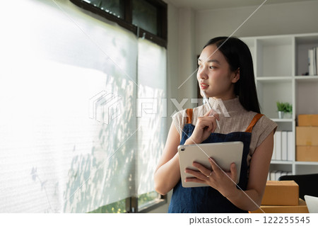 A small SME business owner receives product orders and writes shipping information on tablet in the home office to prepare for delivery. 122255545