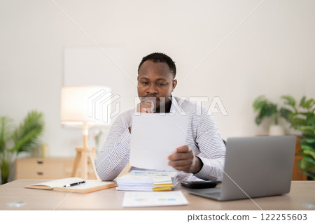 African American businessman feeling stressed while working on laptop, taking notes on documents in office and preparing work report. 122255603
