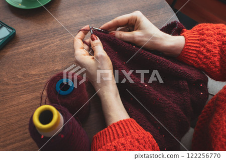 Women hands holding knitting needles and red knitted sweater with sequins. Knitting woman with bobbins of yarn and burgundy knitwear. Sequin pattern in needlework. Knitting hobby. Knitter work.  122256770