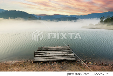 Misty morning view of a rustic wooden pier on a calm lake with a mountainous backdrop Misty morning view of a rustic wooden pier on a calm lake with a mountainous backdrop 122257930