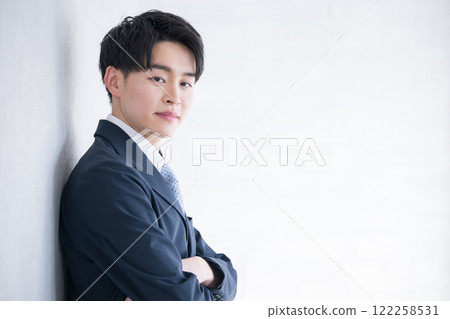Handsome man in his 20s leaning against a wall, wearing a suit, looking back with his arms folded, smiling at the camera; new graduates looking for work or job hunting 122258531
