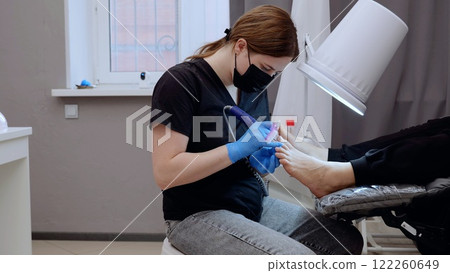 Nail master in a beauty salon giving a professional electric pedicure to a client, using hardware and wearing protective gear 122260649