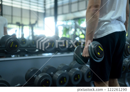 Muscular man holding a pair of dumbbells standing in front of a rack filled with weights at gym Muscular man holding a pair of dumbbells standing in front of a rack filled with weights at gym 122261290