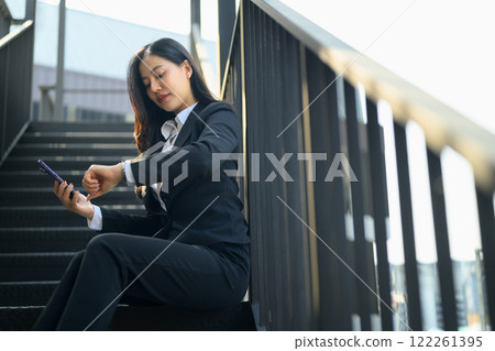 Businesswoman in a suit checking the time on her watch while using phone on the stairs in the city 122261395