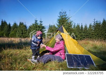 Young child and mother play with smartphone while charging with photovoltaic solar panel near tourist tent in summer. Integration of renewable energy in outdoor camping activities. 122263795