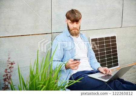 Bearded man works on laptop on lap and smartphone. Solar panel nearby, emphasizing sustainable, modern eco-friendly workspace that combines technology and sustainability. 122263812