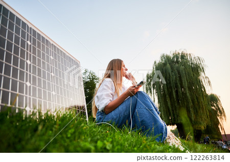 Happy woman using smartphone connected to photovoltaic solar panel. Integration of sustainable renewable energy into everyday life, demonstrating practical use of solar power for charging devices. 122263814