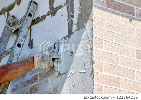 Construction worker applies an even layer of adhesive cement mortar to exterior wall of house while cladding facade with faux brick panels. 122264219