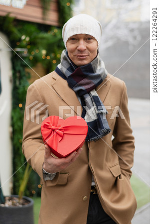 Handsome happy man gives heart-shaped red gift in beige coat on Valentine's Day. Close up. Outdoors portrait in city. Valentine card. 122264291