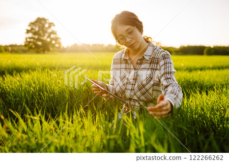 Young farmer woman with digital tablet evaluates shoots, green wheat sprouts in field. Farm work. 122266262