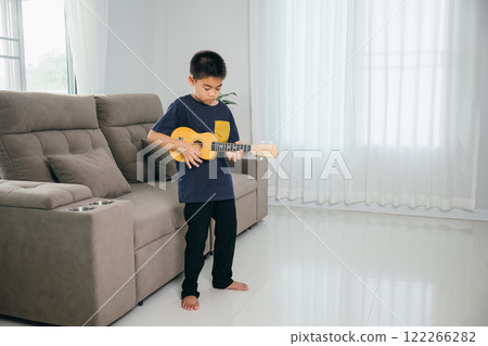 Young boy in casual clothes stands in the living room, playing a ukulele. The soft lighting and homey background accentuate his love for music and creativity. Young boy in casual clothes stands in the living room, playing a ukulele. The soft lighting and homey background accentuate his love for music and creativity. 122266282