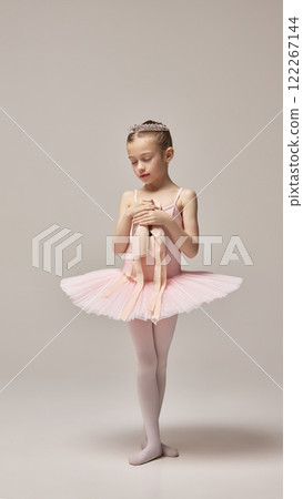 Little ballerina, wearing pink tutu and tiara, standing calmly, with closed eyes, holding pointe shoes in hands against white studio background. 122267144