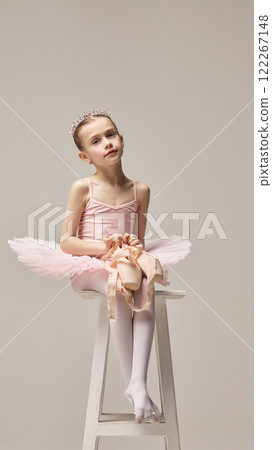 Charming little ballerina, wearing pink tutu and tiara, sitting gracefully on stool holding pointe shoes with calm expression against white background. 122267148
