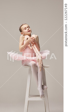 Joyful child, ballerina sitting on stool holding pointe shoes close to chest wearing pink tutu and tiara against white studio background. 122267149