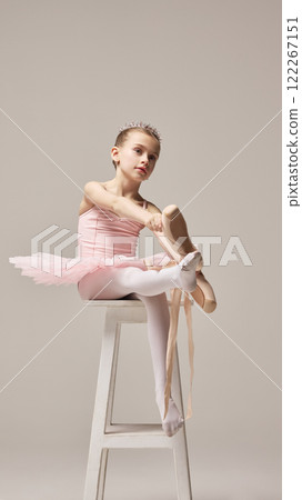 Charming little ballerina seated on white stool, adjusting pointe shoes, wearing tiara and pink tutu, looks thoughtful against warm grey background. Charming little ballerina seated on white stool, adjusting pointe shoes, wearing tiara and pink tutu, looks thoughtful against warm grey background. 122267151