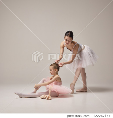 Girl in pink ballet outfit seated adjusting ribbons on pointe shoes, adult ballerina standing nearby offering guidance against light background. 122267154