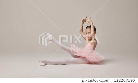 Little girl, ballerina dancer in tutu performs sitting pose with one leg extended, arms elegantly positioned, expressing charm and skill against beige background. Little girl, ballerina dancer in tutu performs sitting pose with one leg extended, arms elegantly positioned, expressing charm and skill against beige background. 122267180