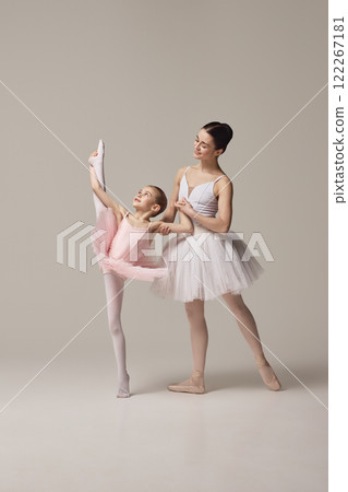 Ballerina in white tutu supports little dancer in pink tutu performing leg stretch, emphasizing guidance and flexibility against beige studio background. Ballerina in white tutu supports little dancer in pink tutu performing leg stretch, emphasizing guidance and flexibility against beige studio background. 122267181
