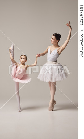 Ballerina in white tutu supports little girl, dancer in pink tutu performing leg stretch, emphasizing guidance and flexibility against white background. Ballerina in white tutu supports little girl, dancer in pink tutu performing leg stretch, emphasizing guidance and flexibility against white background. 122267183