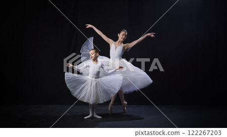 Little kid and ballerina in white tutus gracefully perform synchronized arm movements, displaying elegance and poise against black studio background. 122267203