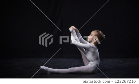 Young ballerina holding one leg extended high with arms, displaying balance and flexibility, dressed in white suit, against black studio background. Young ballerina holding one leg extended high with arms, displaying balance and flexibility, dressed in white suit, against black studio background. 122267210