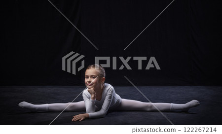Kid, ballerina smiling while holding forward split, hands on chin, dressed in white, expressing flexibility and poise, against black studio background. Kid, ballerina smiling while holding forward split, hands on chin, dressed in white, expressing flexibility and poise, against black studio background. 122267214