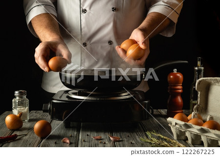 In a cozy kitchen, a chef holds two fresh eggs, poised to crack them into a hot skillet. The warm steam rises, hinting at a savory dish in the making, surrounded by rustic ingredients In a cozy kitchen, a chef holds two fresh eggs, poised to crack them into a hot skillet. The warm steam rises, hinting at a savory dish in the making, surrounded by rustic ingredients 122267225