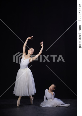 Adult ballerina posing with arms elegantly curved above head, child sitting nearby gazing upwards, both in white costumes, against black studio background. 122267246